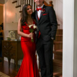 A young couple dressed formally, with the woman in a long red gown holding flowers and the man in a black tuxedo with a red bow tie, stand smiling together in a warmly lit home near a staircase.