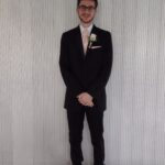 A man wearing a black suit, light-colored tie, and a white boutonnière stands smiling in front of a light gray, vertically-striped wall and tiled floor.