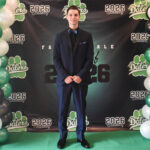A young man in a dark suit stands smiling between black, white, and green balloon columns in front of a backdrop that reads Dalers and 2026, at what appears to be a formal school event.