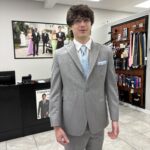 A young man in a light gray suit with a light blue tie and pocket square stands in a formalwear store. Behind him are racks of ties and a large photo of people dressed in formal attire.