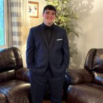 A young man in a navy suit and black shirt stands smiling in a living room with brown leather sofas, a leafy plant, and a window with striped curtains. A framed photo hangs on the light-colored wall behind him.