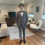 A young man in a gray suit with a bow tie stands smiling in a modern living room with wooden floors, white chairs, a TV, and patterned curtains.