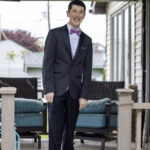 A young man in a dark suit with a purple shirt and bow tie stands and smiles on a porch with outdoor furniture in the background.