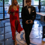 A woman in a red gown holding red flowers walks beside a man in a black tuxedo inside an elegant venue with large windows, marble floors, and decorative patterns.