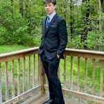 A young man in a black suit with a blue patterned tie stands on a wooden deck, hands in pockets, with green trees and grass in the background.