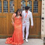 Two teenagers stand on stone steps in front of ornate wooden doors. The girl wears a long, orange sequin dress, and the boy wears a white suit with an orange tie. They both look down towards the ground.