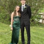 A young couple dressed for prom stands on a grassy lawn. The girl wears a green satin dress and corsage, while the boy wears a black suit with a green tie and boutonniere. Green shrubbery and a wooden fence are in the background.