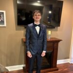 A young man in a dark patterned tuxedo with a black bow tie stands smiling in front of a fireplace and TV in a warmly lit living room with hardwood floors and tan walls.