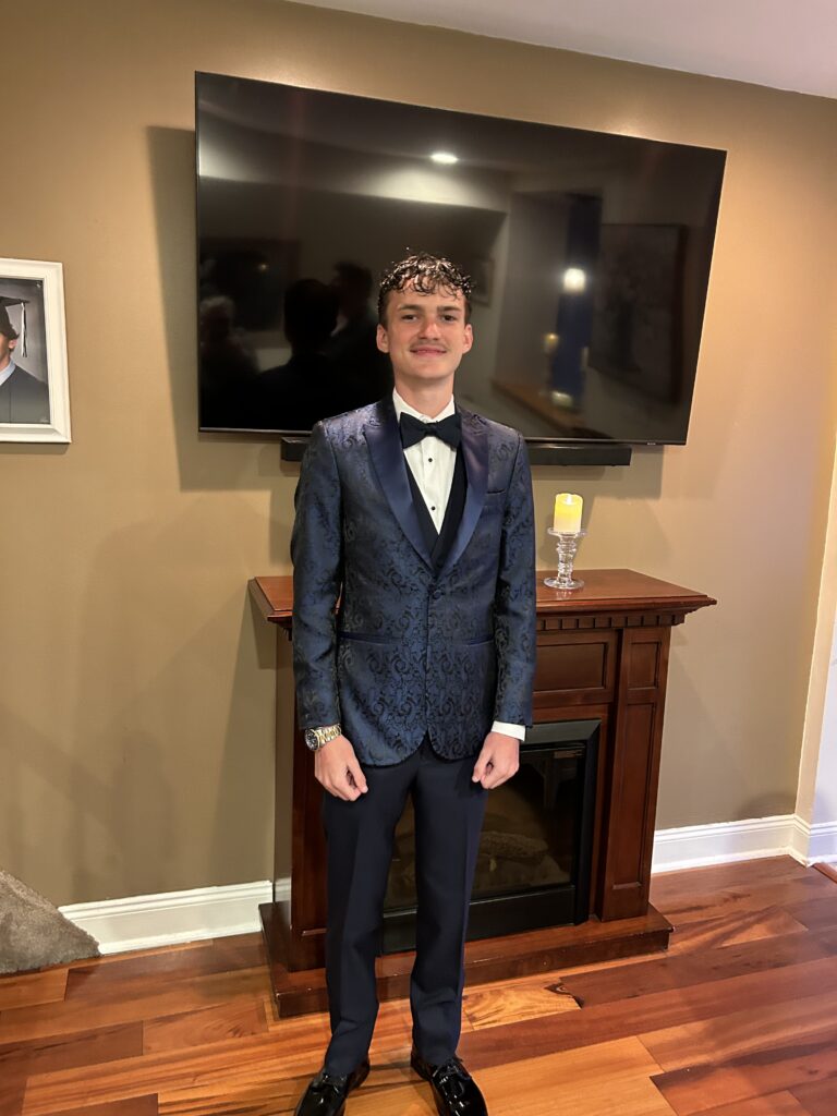A young man in a dark patterned tuxedo with a black bow tie stands smiling in front of a fireplace and TV in a warmly lit living room with hardwood floors and tan walls.