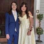 Two teenagers dressed for a formal event stand side by side. The person on the left wears a blue suit and glasses, while the person on the right wears a light yellow, off-shoulder gown with a floral corsage. They smile in front of a wooden door.