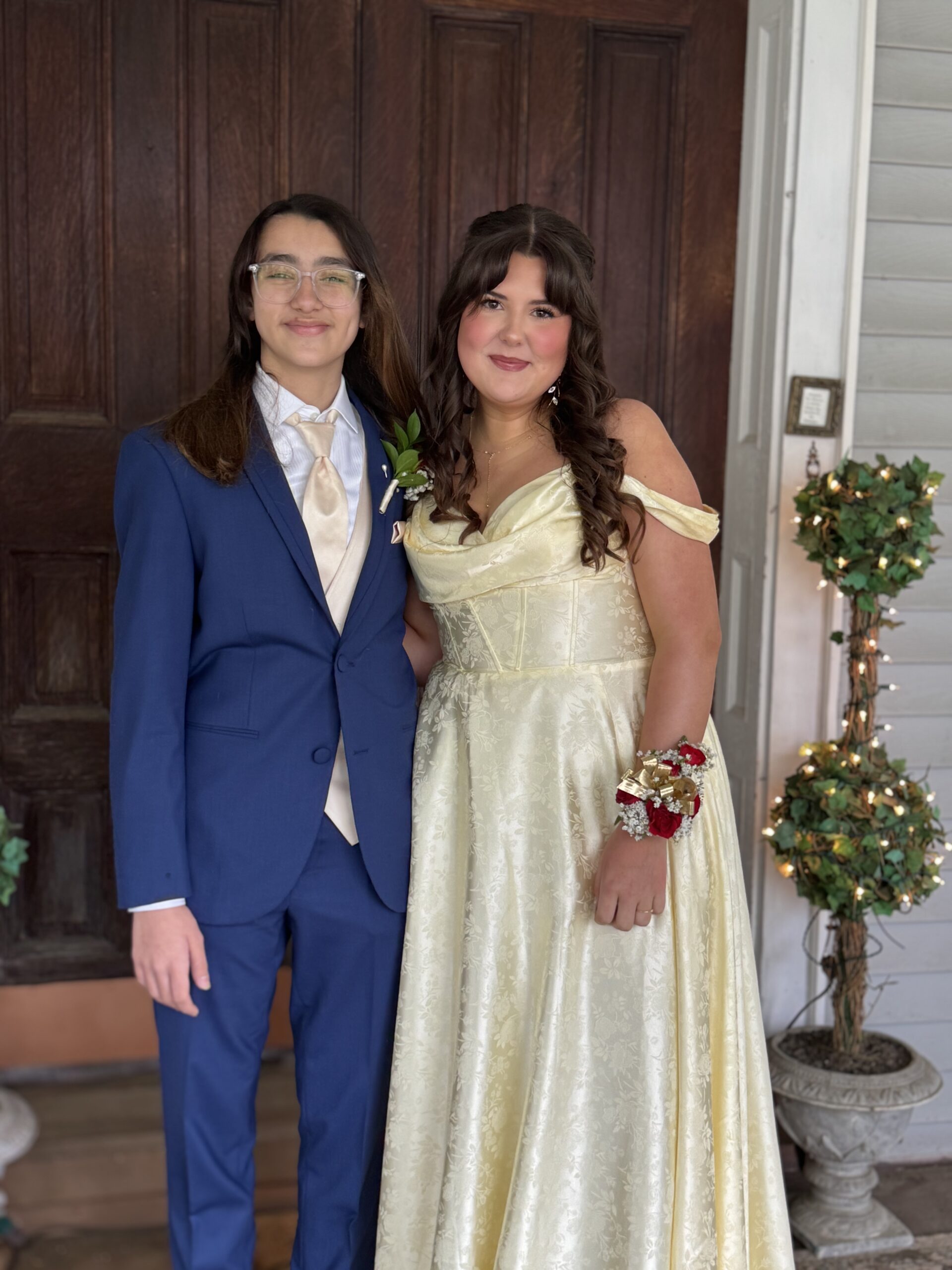 Two teenagers dressed for a formal event stand side by side. The person on the left wears a blue suit and glasses, while the person on the right wears a light yellow, off-shoulder gown with a floral corsage. They smile in front of a wooden door.