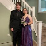 A young man in a black suit and bow tie stands next to a young woman in a sparkly purple gown holding a bouquet, both smiling and posing in front of a staircase inside a house.