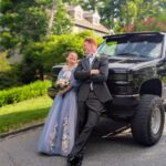 A young couple dressed in formal attire stands smiling and leaning against a black pickup truck on a sunny day, with trees, greenery, and a house in the background.