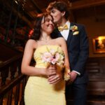 A young couple dressed formally stands on a staircase. The woman in a yellow dress holds a bouquet and smiles, while the man in a suit kisses her on the forehead, creating a warm, affectionate moment.
