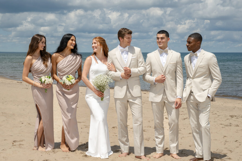 Five people dressed in formal attire stand barefoot on a sandy beach, smiling and talking. Three women wear light dresses and hold flowers, while two men wear light suits with bow ties. The ocean and cloudy sky are in the background.Best Suit Styles for Summer Weddings on the East Coast