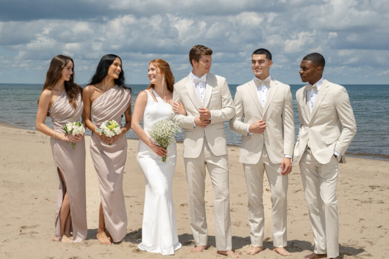 Five people dressed in formal attire stand barefoot on a sandy beach, smiling and talking. Three women wear light dresses and hold flowers, while two men wear light suits with bow ties. The ocean and cloudy sky are in the background.Best Suit Styles for Summer Weddings on the East Coast