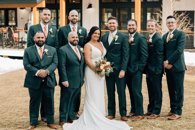 A bride in a white dress holding a bouquet stands outside with eight groomsmen in dark green suits with orange ties, all smiling and posing for a wedding photo on a lawn in front of a building. Formalwear for Every Body Type: Why In-Person Fittings Matter