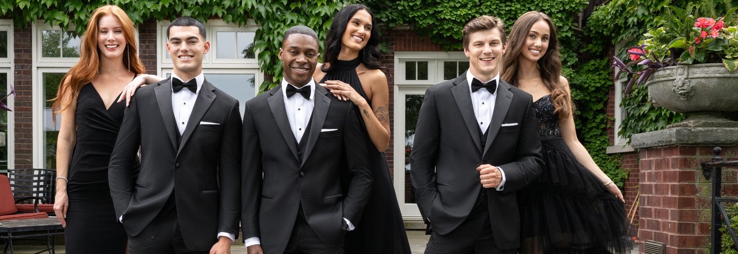 Six young adults dressed in formalwear pose together outdoors; three men in black tuxedos stand in front, while three women in elegant black dresses stand behind them, all smiling and standing by a building covered in ivy.