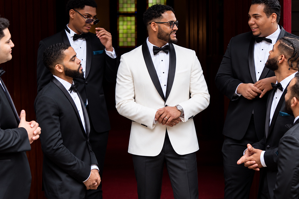 Groom in white wedding dinner jacket standing with groomsmen in black tuxedos by Tuxedo By Sarno.
